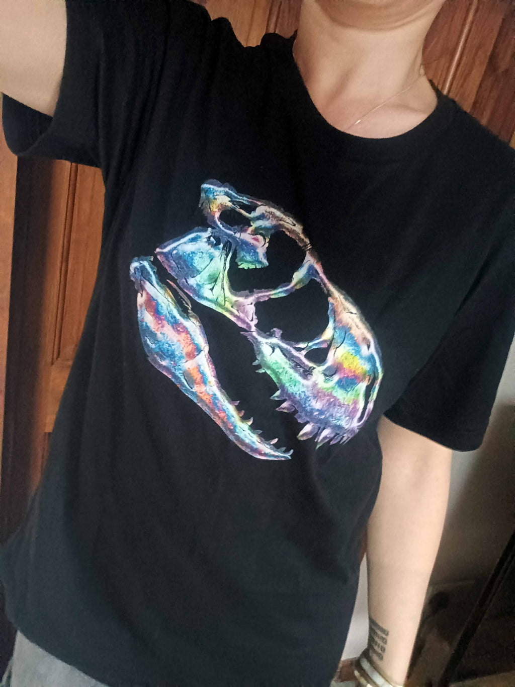 A person wearing a black short-sleeve T-shirt with a large colorful petrol spill inspired T-rex skull graphic on the front, photographed indoors against a wooden door background.