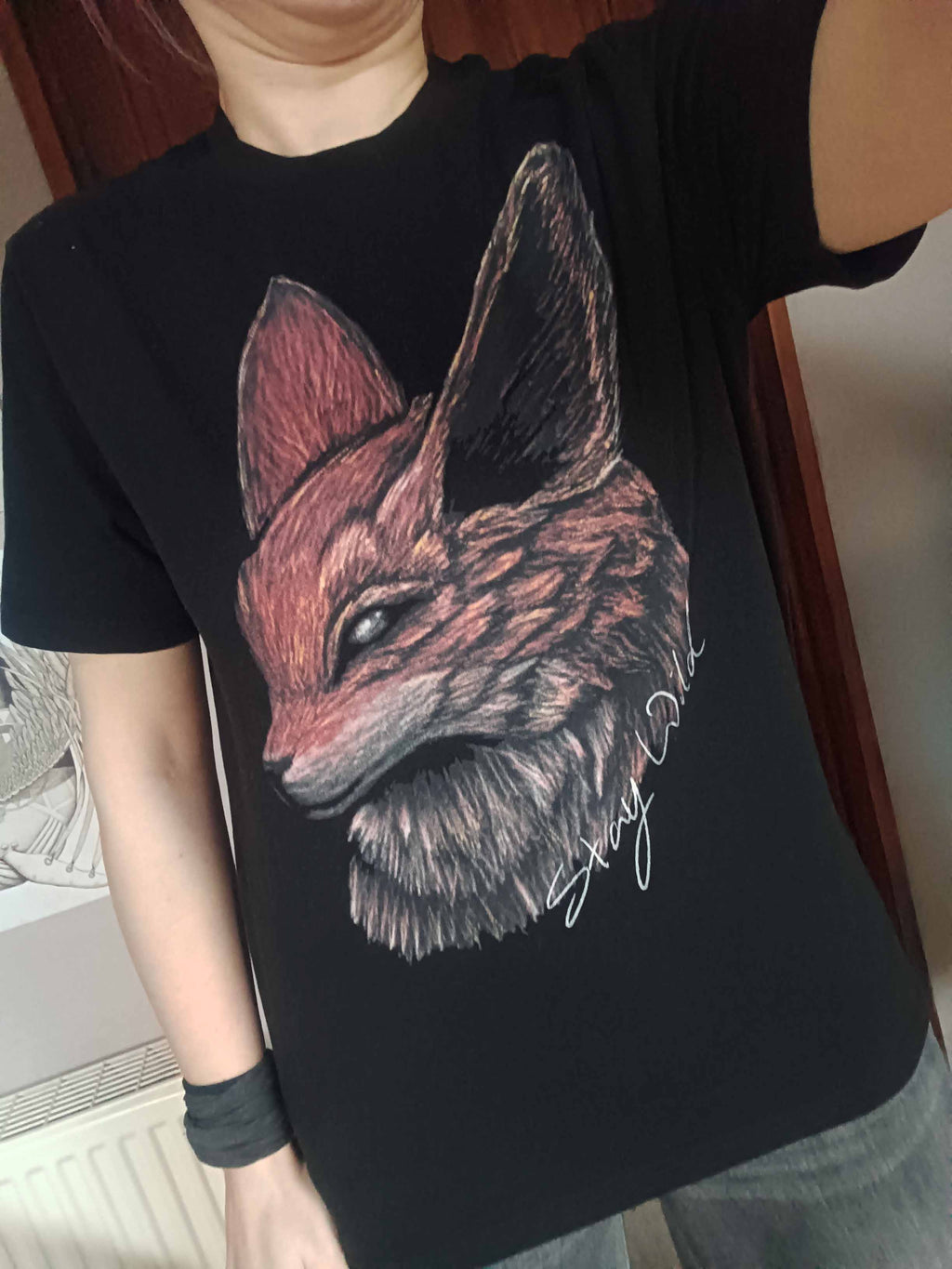 A person wearing a black short-sleeve T-shirt with a large illustrated fox head graphic and the words "stay wild" in white on the front, photographed indoors.