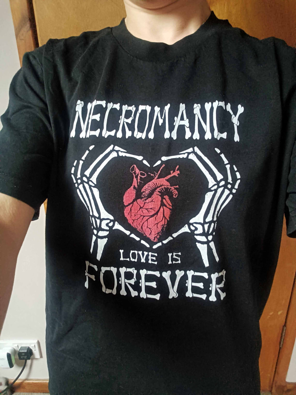 A person wearing a black short-sleeve graphic t-shirt with a white skeletal heart design and a red anatomical heart in the center, with the text 'NECROMANCY' and 'LOVE IS FOREVER'.