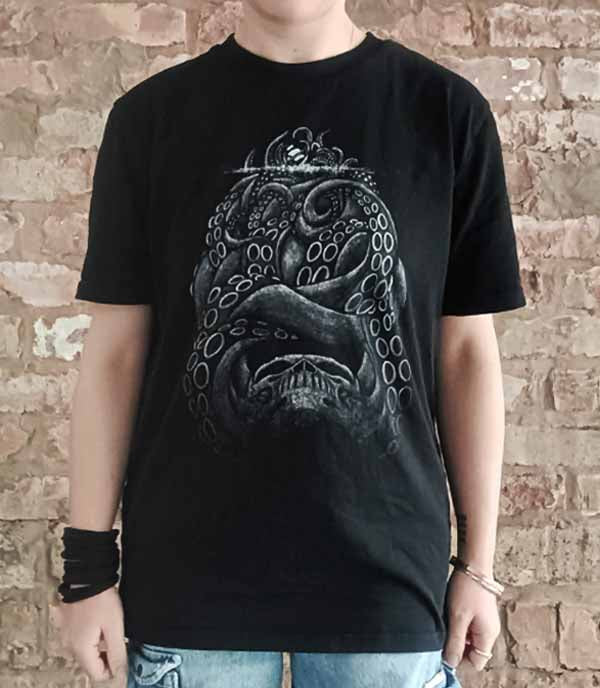 Person wearing a black t-shirt with a detailed design  of a kraken attacking a ship in front of a brick wall.