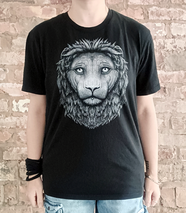 A person wearing a black short-sleeve crewneck t-shirt with a large gray line art graphic of a lion's face on the front, standing against a rustic brick wall.