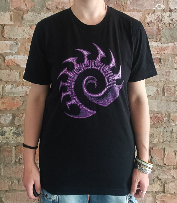 A person wearing a black short-sleeve t-shirt with a large purple zerg logo graphic on the front, standing against a brick wall background.