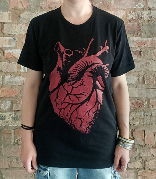 A person wearing a black short-sleeve graphic T-shirt with a large distressed red anatomical heart print on the front, paired with light blue jeans, standing against a rustic brick wall.