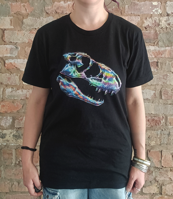 A person wearing a black short-sleeve T-shirt with a large colorful petrol spill inspired T-rex dinosaur skull graphic on the front, standing against a rustic brick wall.