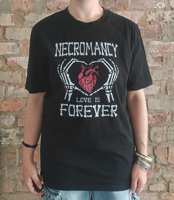 A person wearing a black graphic t-shirt with a white skeleton hand holding a red anatomical heart and the text 'NECROMANCY LOVE IS FOREVER', standing against a brick wall.