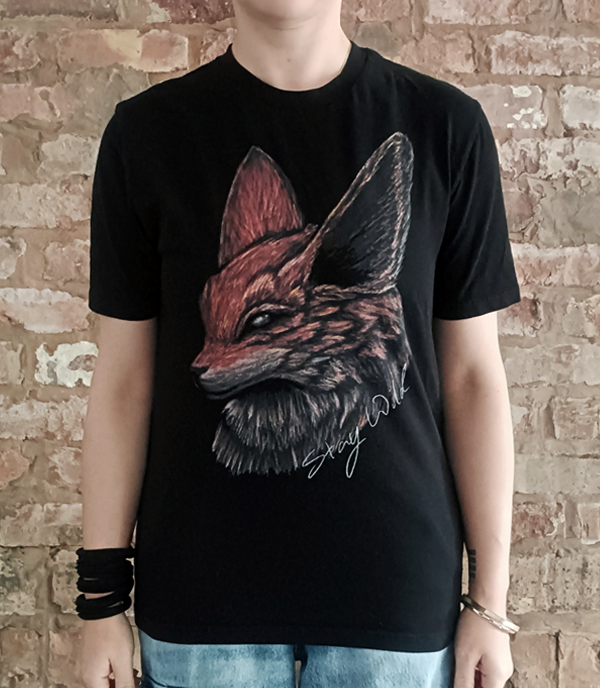 A person wearing a black short-sleeve t-shirt with a large graphic print of a fox head in profile, featuring red, brown, and gray tones with the words "stay wild" in  white handwriting. The person is standing in front of a rustic brick wall.
